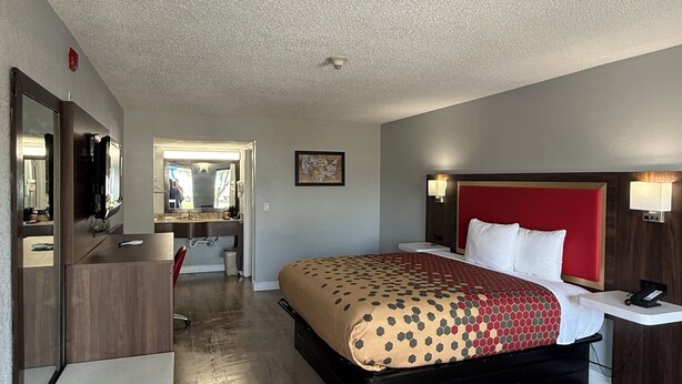 A hotel room with a bed, a desk, a chair, a mirror, and a nightstand with a lamp.