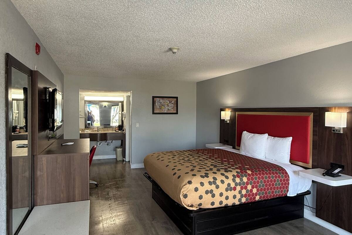 A hotel room with a bed, a desk, a chair, a mirror, and a nightstand with a lamp.
