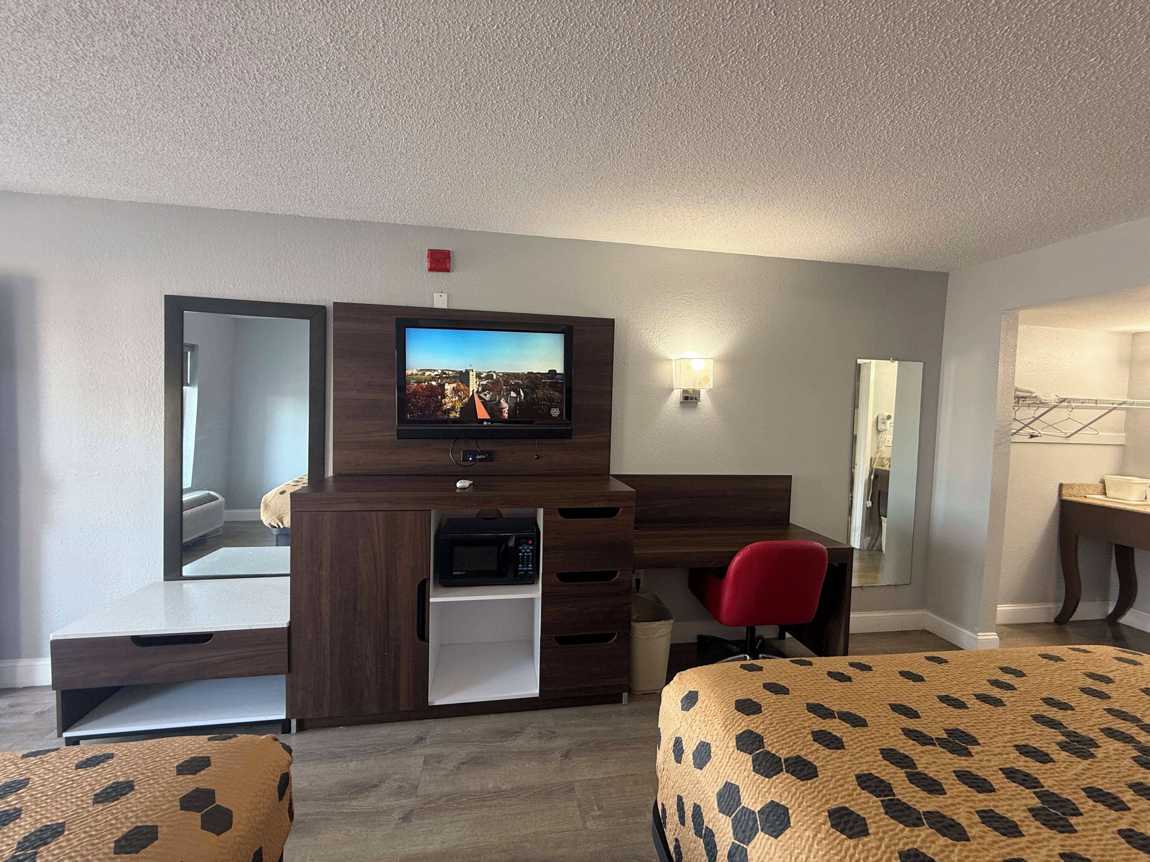 Une chambre d’hôtel équipée d’une télévision, d’un bureau, d’un four à micro-ondes, d’une chaise rouge et d’un lit recouvert d’une couverture à motifs.