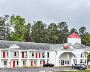 Un motel composé d’un bâtiment principal doté d’un toit rouge et d’une façade blanche, de nombreuses portes rouges et d’un parking.