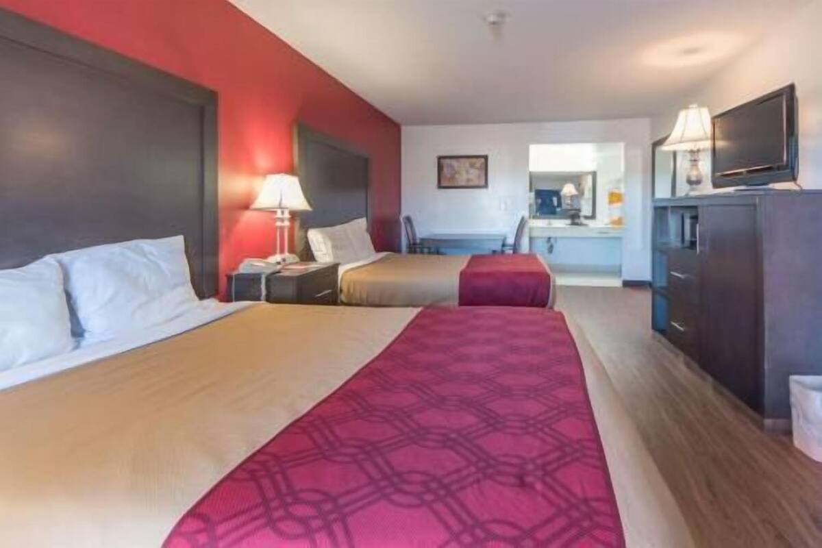 A hotel room with two beds, a desk, a chair, a TV, and a lamp.