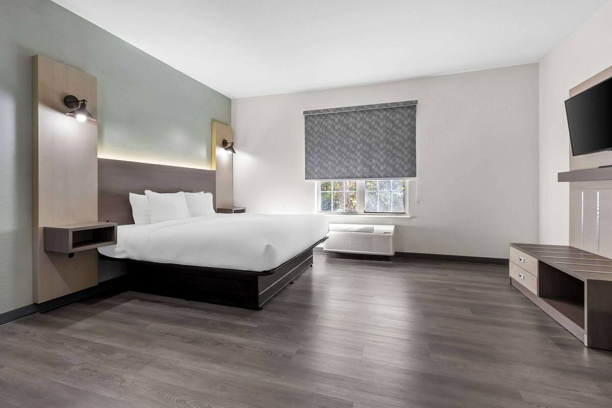 A modern bedroom with a large bed, built-in nightstands, a flat-screen TV, and a window with blinds.