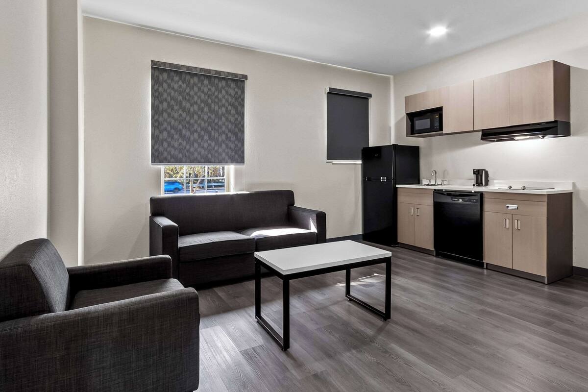 A compact living space with a sofa, a coffee table, a kitchen area including a microwave and dishwasher, and a window with blinds.