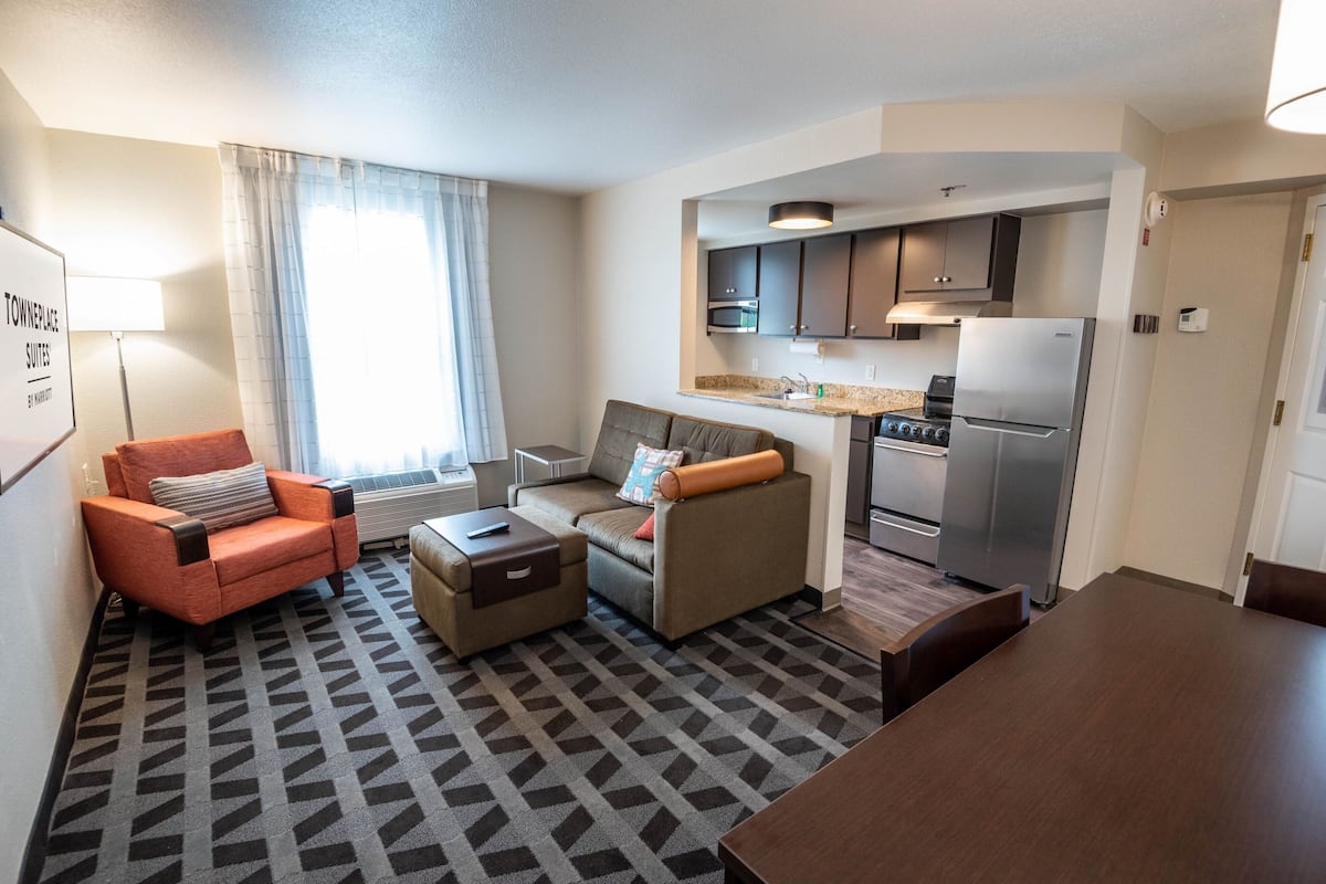Un espace de vie compact comprenant un canapé, un fauteuil et un pouf, une kitchenette avec un réfrigérateur et une cuisinière, ainsi qu’une table à manger.