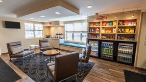 A seating area with sofas and a table, a snack bar with shelves of snacks and drinks, and a window with a view of the parking lot. - TownePlace Suites by Marriott Gahanna (Columbus)