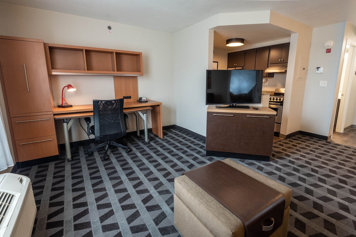 Une chambre d’hôtel compacte comprenant un coin cuisine, un bureau avec une chaise, une télévision et une moquette à motifs.