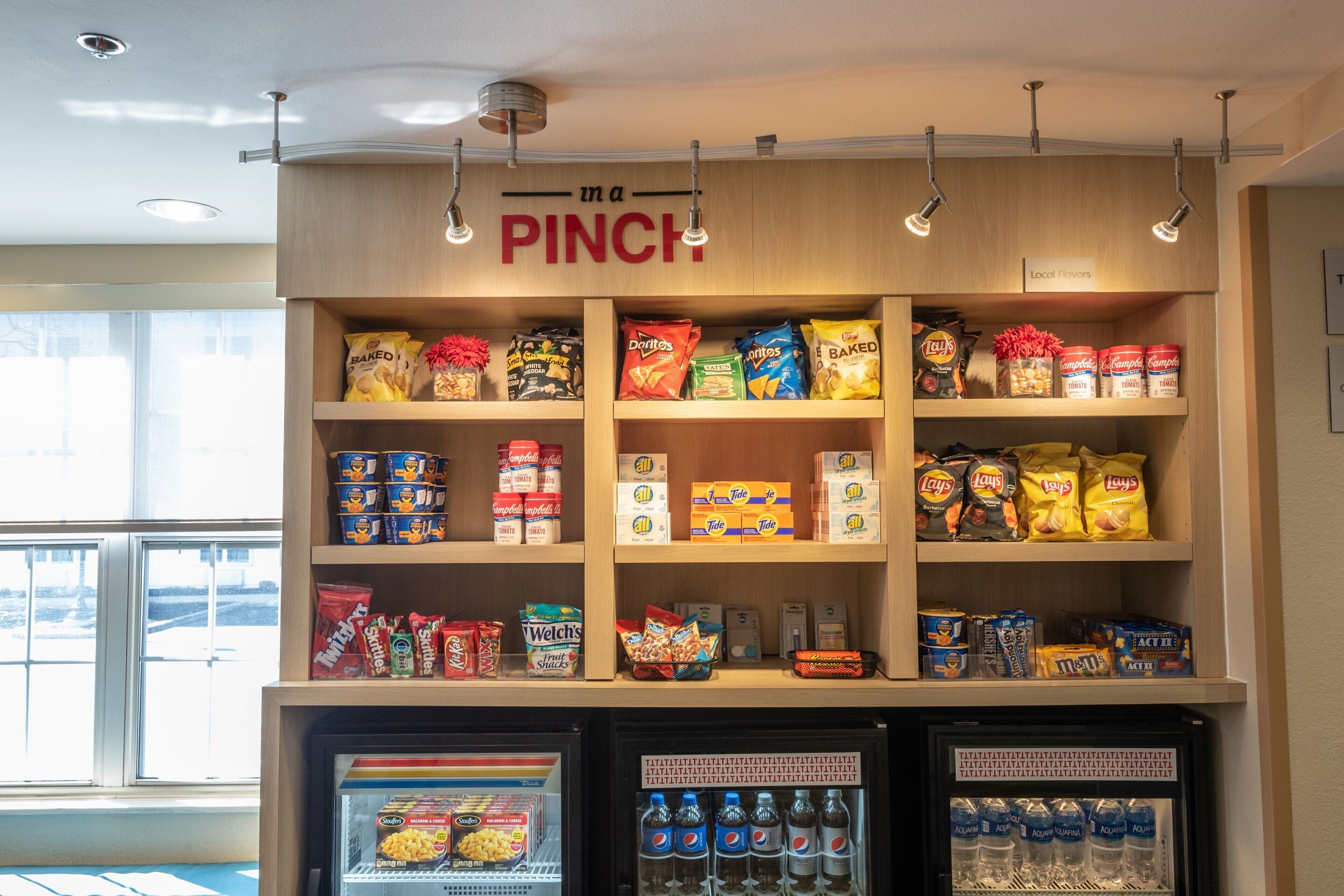 Un présentoir proposant divers types de chips et d’en-cas, un rayon réfrigéré avec des boissons, et une pancarte au-dessus indiquant : « en cas d’urgence ».