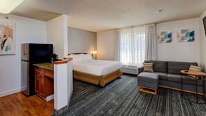 Habitación de hotel con cama, sofá, una mesita, televisor, zona de cocina y obras de arte en las paredes.