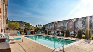 Un área de piscina con sillones de descanso, una piscina cristalina y un edificio al fondo con fachadas de ladrillo y piedra.