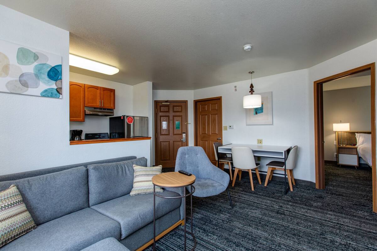 A modern hotel room with a grey sofa, a small round table, a dining area with a white table and chairs, wooden cabinets, and a kitchen area with a refrigerator and coffee machine.