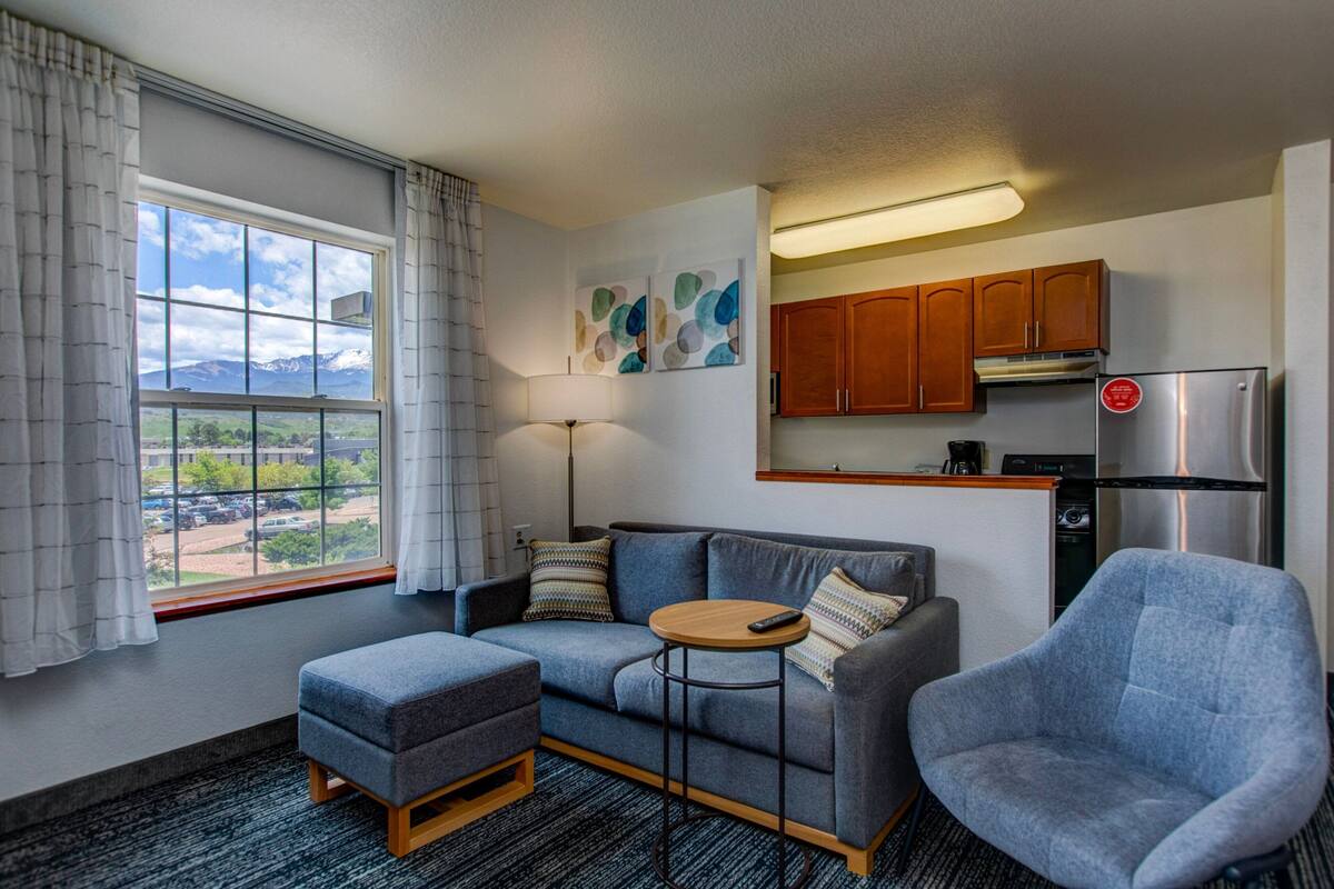 A compact living space with a sofa, ottoman, and armchair, a small round table, a kitchenette with wooden cabinets, and a window with a view of mountains and a parking lot.