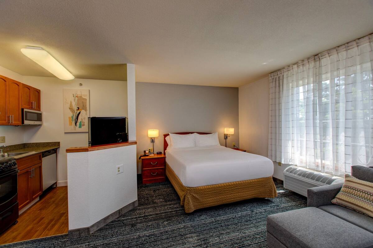 A hotel room with a kitchenette, a bed, a TV, and a large window with curtains.