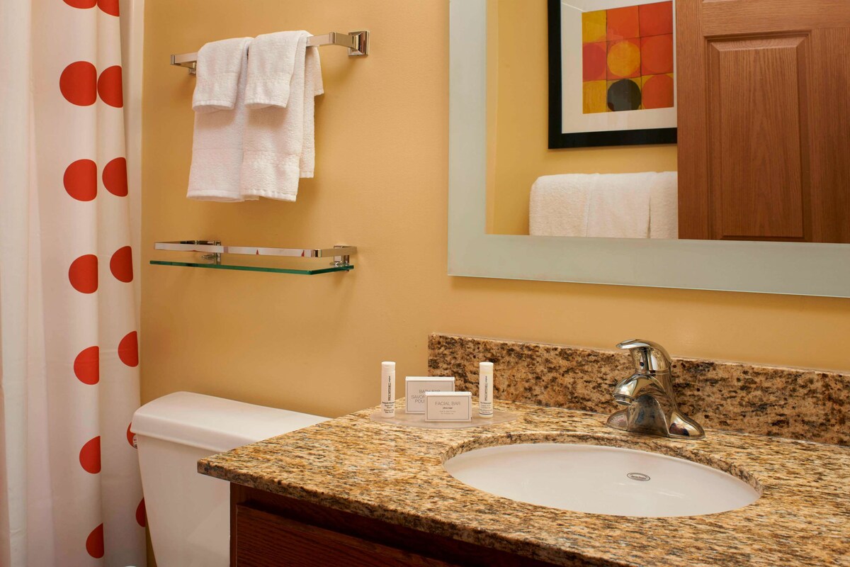 A bathroom with a granite sink, a toilet, a shower curtain with red polka dots, a towel rack with towels, a glass shelf, and a framed artwork on the wall.