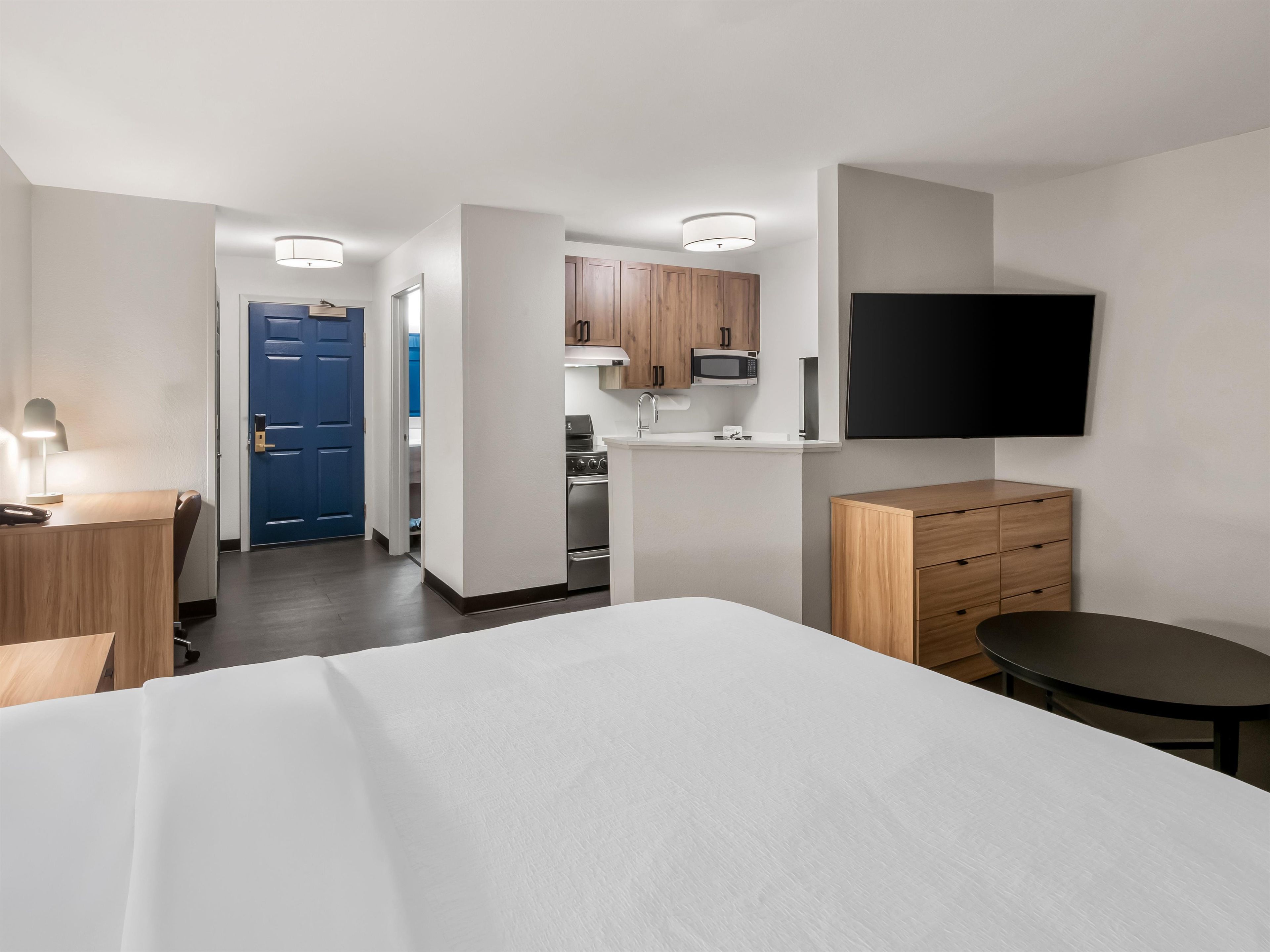 A hotel room with a bed, desk, chair, TV, and kitchenette.