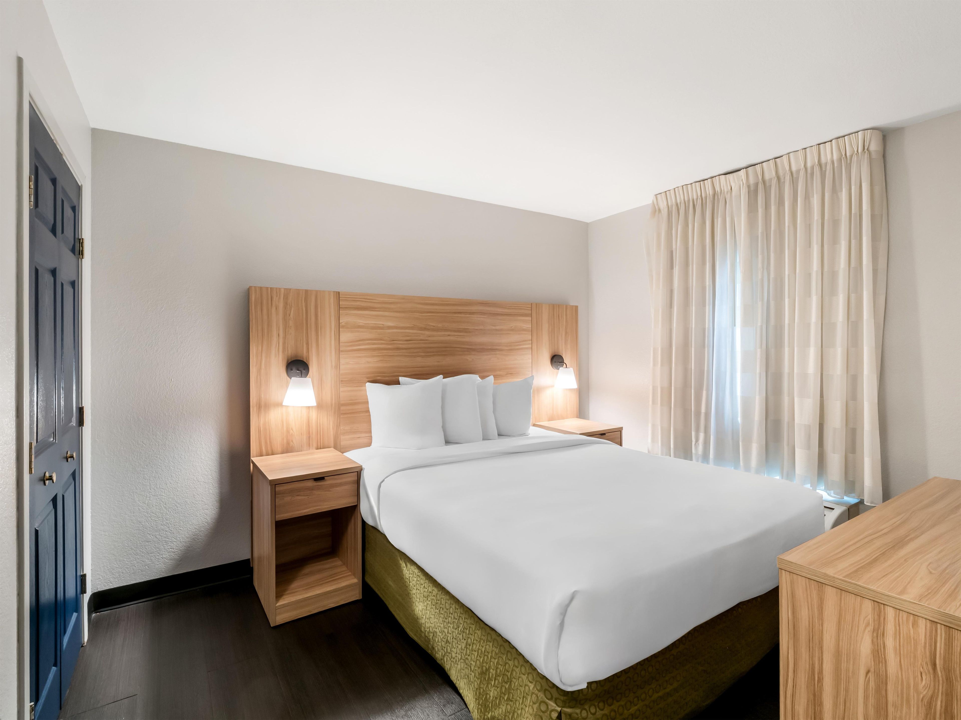 A hotel room with a wooden headboard, a bed with white linens, two bedside lamps, and a window with sheer curtains.