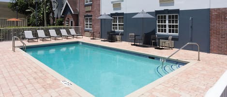 Une piscine rectangulaire bordée de carreaux, entourée de chaises longues et de parasols, et située à côté d’un bâtiment à la façade en briques et aux grandes fenêtres.