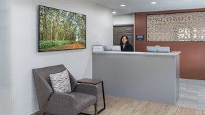 Uma área de recepção com um balcão moderno, uma cadeira com almofada estampada e uma grande moldura com uma imagem de uma floresta na parede.