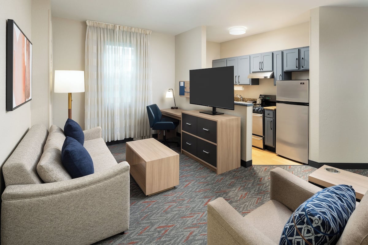A hotel room with a sofa, a desk, a television, a kitchenette, and a dining table.