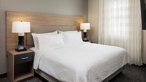 Une chambre d’hôtel avec un grand lit, deux tables de chevet avec des lampes, une petite table de nuit avec un tiroir et une fenêtre avec des rideaux.