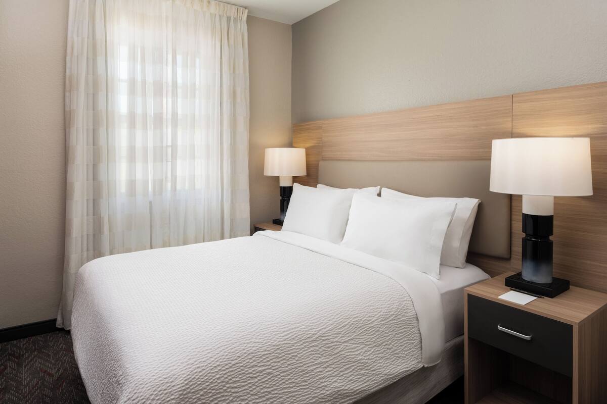 A hotel room with a large bed, two bedside tables with lamps, a wooden headboard, and sheer curtains.