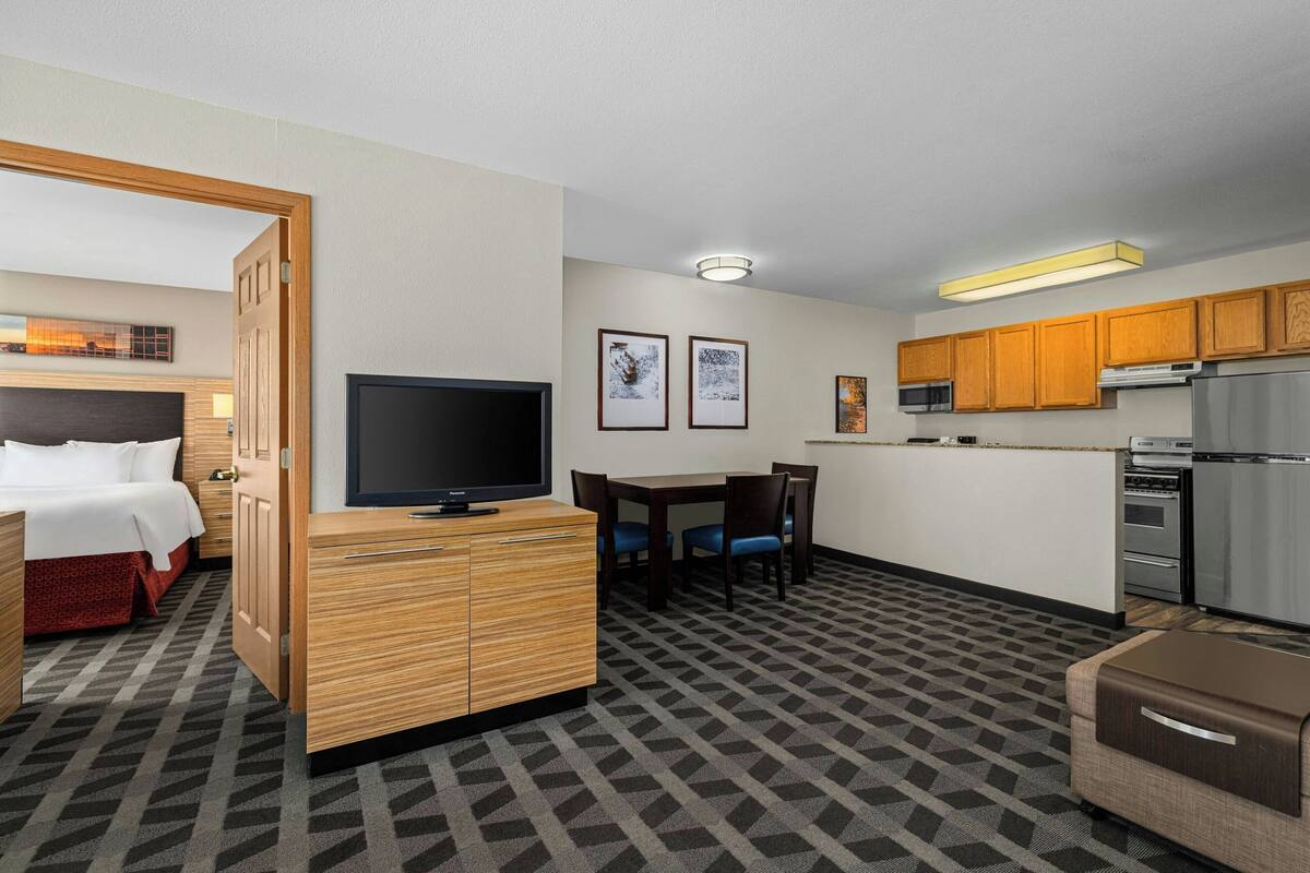 A hotel room with a bed, TV, wooden cabinet, dining table with chairs, kitchen area with cabinets and appliances, and patterned carpet.