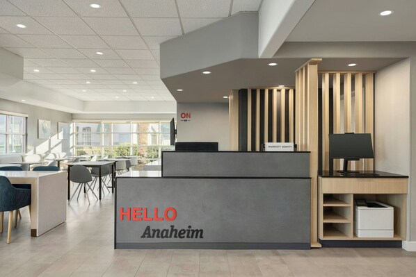 A modern reception desk with a "HELLO Anaheim" sign, a computer, and a seating area with tables and chairs.