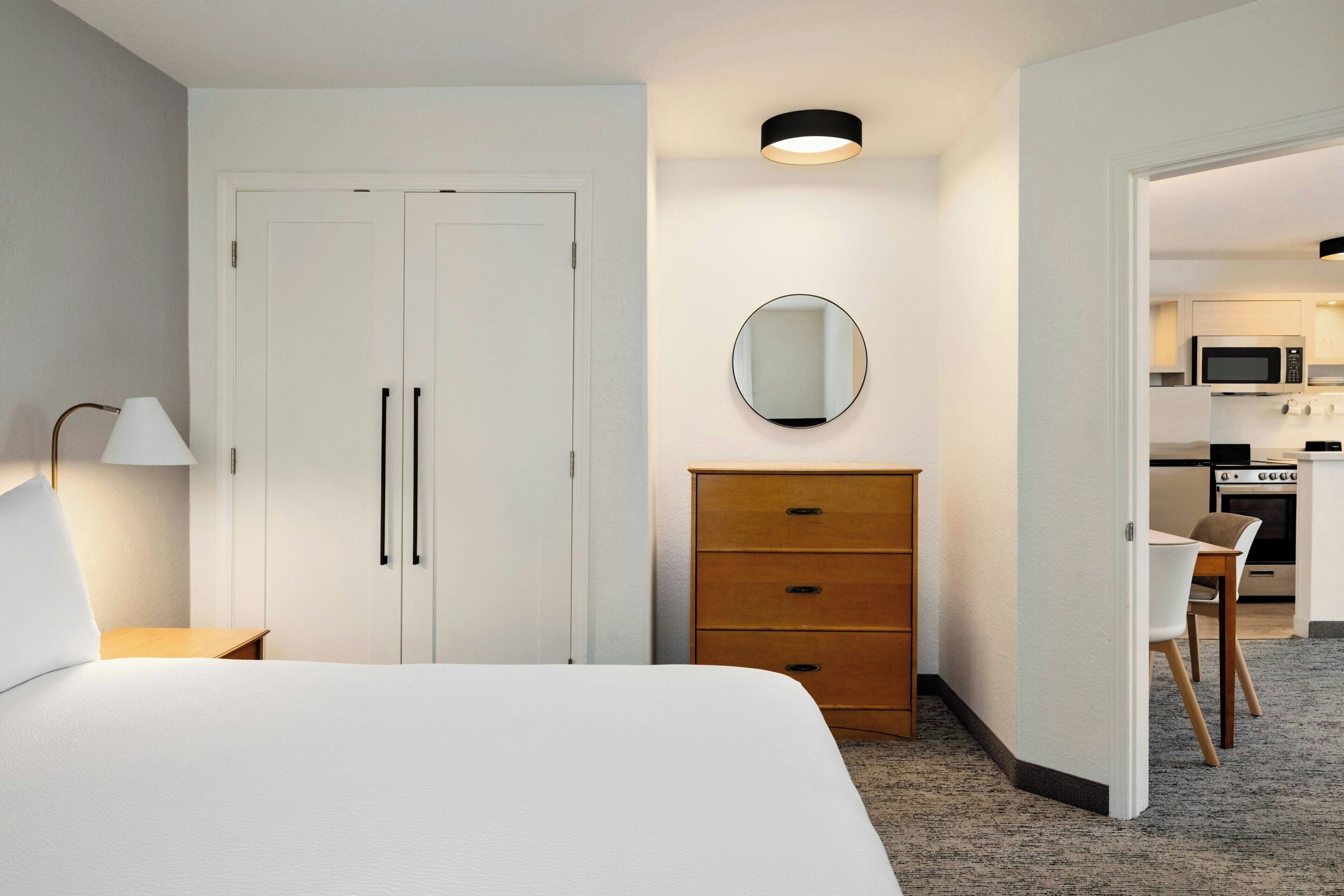 A hotel room with a bed, a dresser, a mirror, a desk, and a kitchenette in the background.