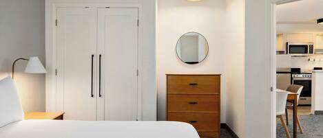 A hotel room with a bed, a dresser, a mirror, a desk, and a kitchenette in the background.