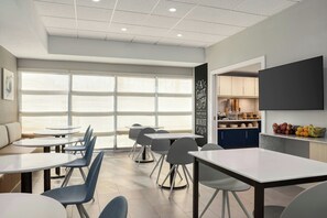 A modern dining area with round tables and chairs, a kitchen counter with fresh produce, and large windows providing natural light.