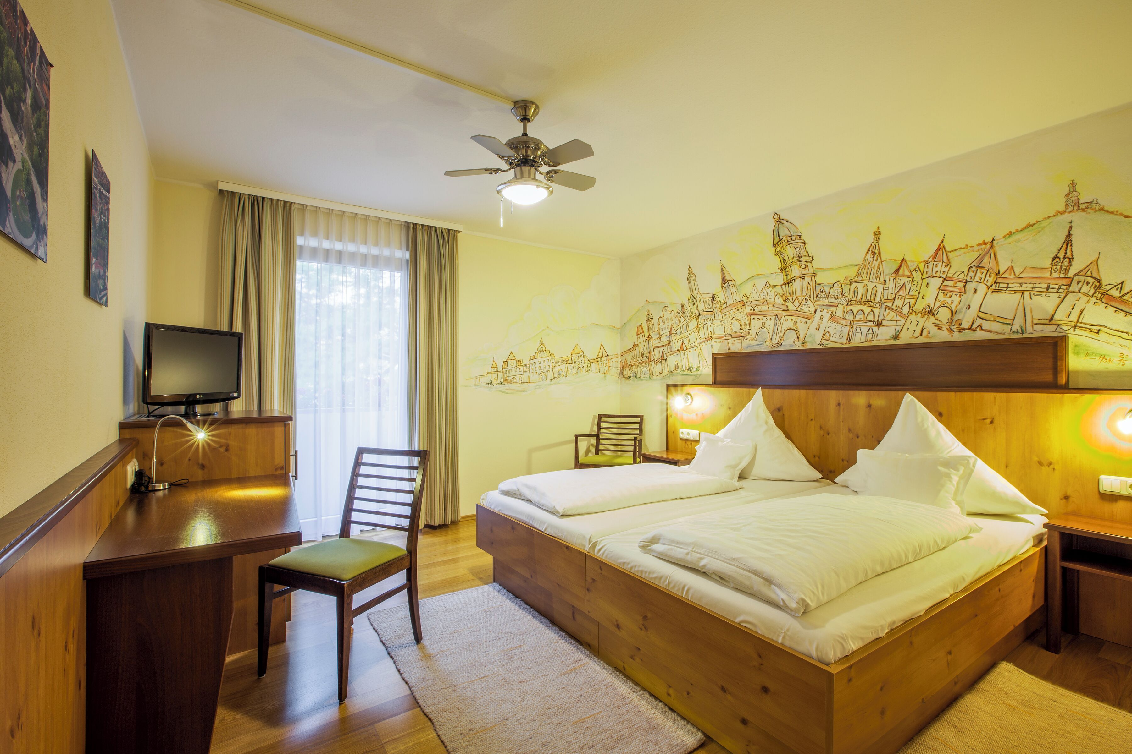 Habitación de hotel con cama, escritorio, silla, televisor y ventilador de techo.