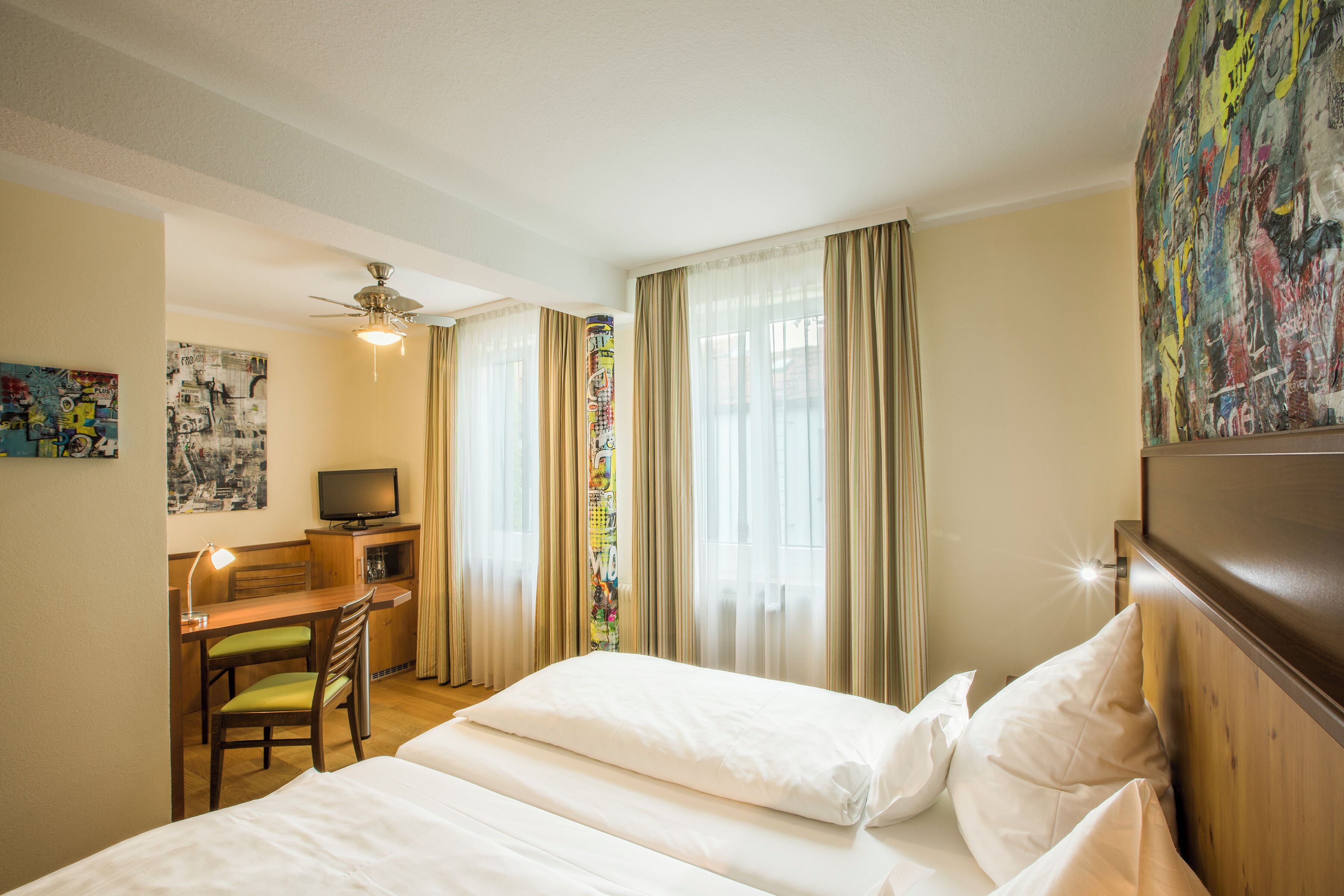 Una habitación de hotel con cama, escritorio con silla, televisión y ventilador de techo.
