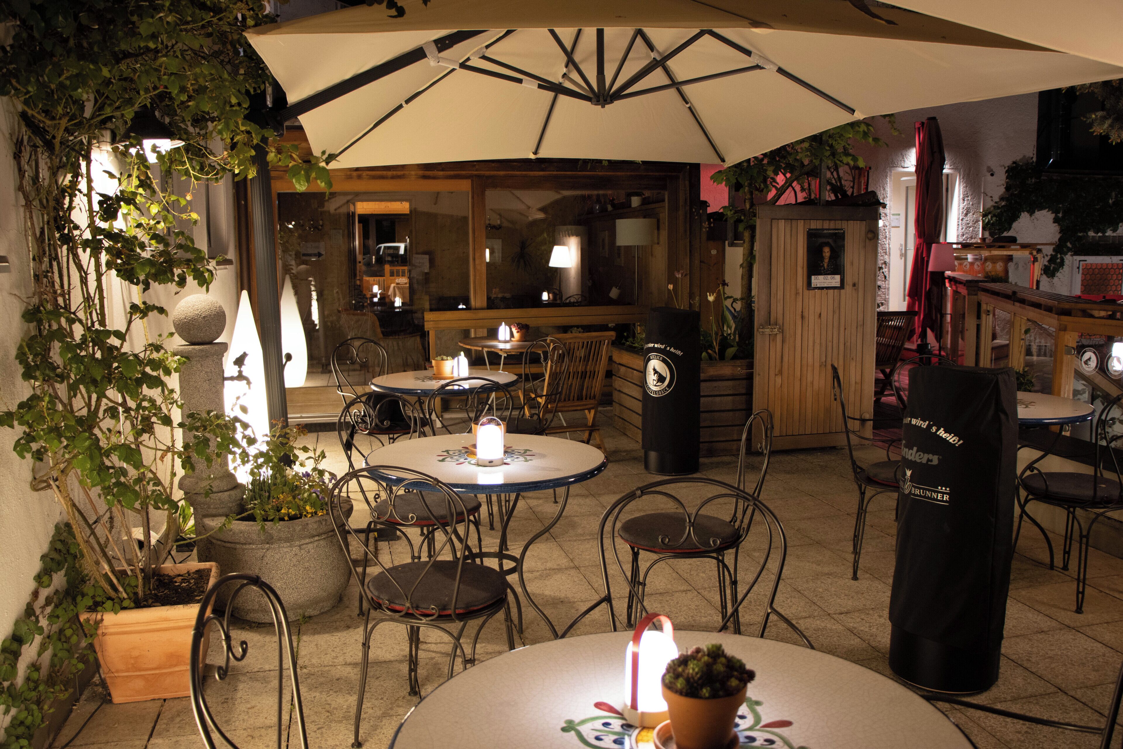 An outdoor seating area with round tables and chairs, a large umbrella providing shade, and potted plants.