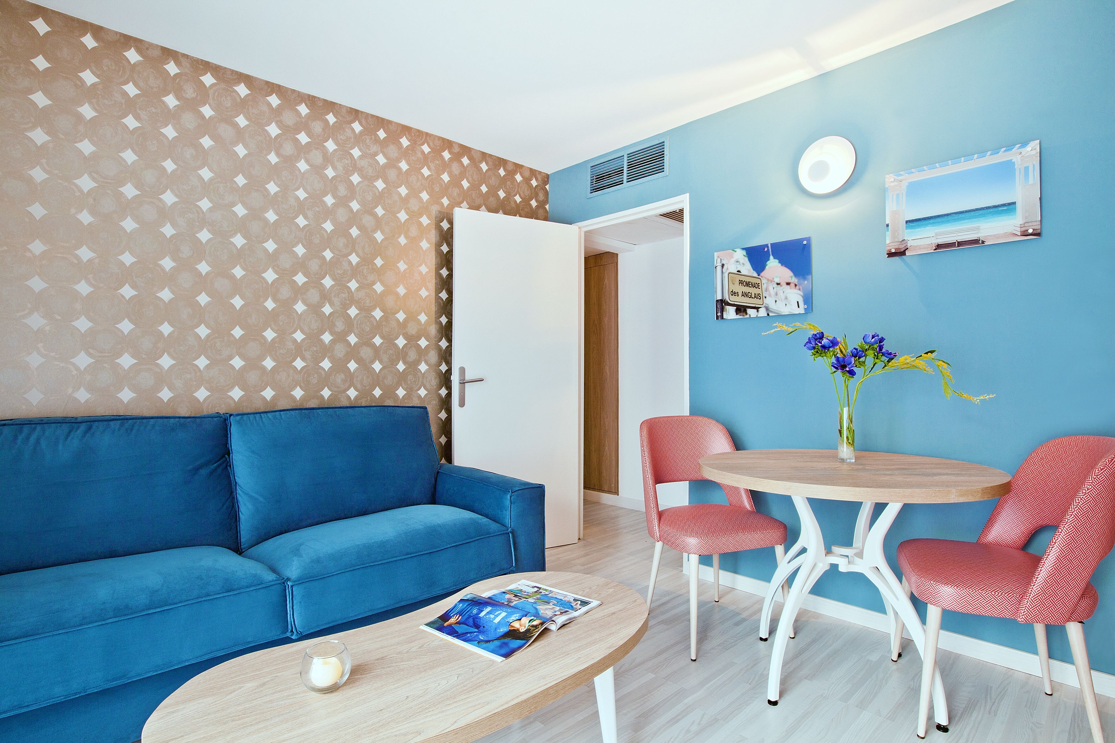 Ein Zimmer mit einem blauen Sofa, einem runden Holztisch mit zwei roten Stühlen, einer kleinen Vase mit Blumen und einer Wand mit blau-weißem Muster.