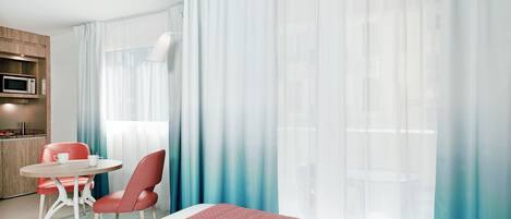 A hotel room with a bed, a dining table, chairs, a microwave, and sheer curtains.