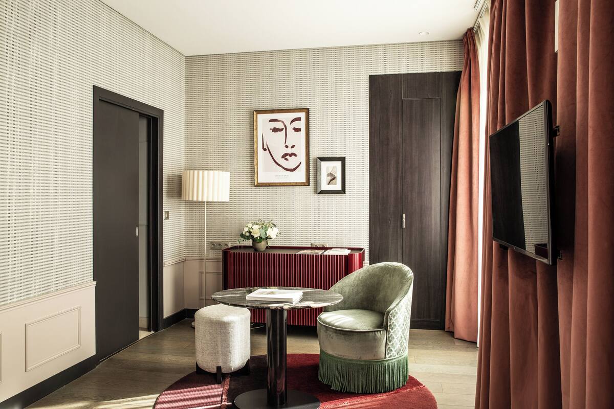 A modern hotel room with a large window, a flat-screen TV, a round table, a comfortable armchair, and a decorative wall piece.