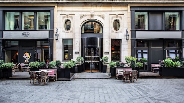 A hotel entrance with a black revolving door, outdoor seating with wicker chairs and tables, and a cobblestone pavement. - Hotel Indigo Paris - Opera by IHG (Paris)