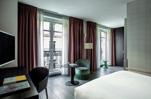 A hotel room with a bed, a desk with a chair, a small table with a chair, a lamp, and a view of the outside. - Hotel Indigo Paris - Opera by IHG (Paris)