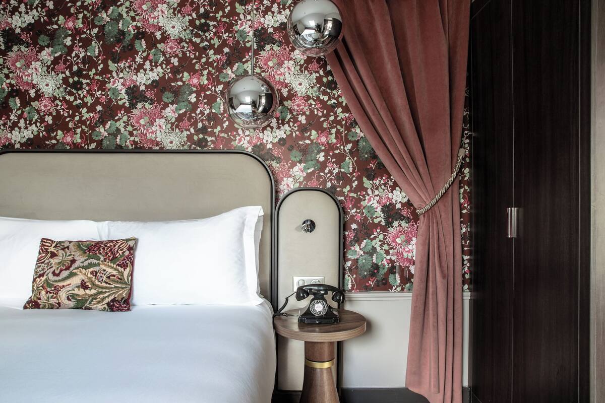 A bed with a floral-patterned pillow, a bedside telephone, and a wall with floral wallpaper.