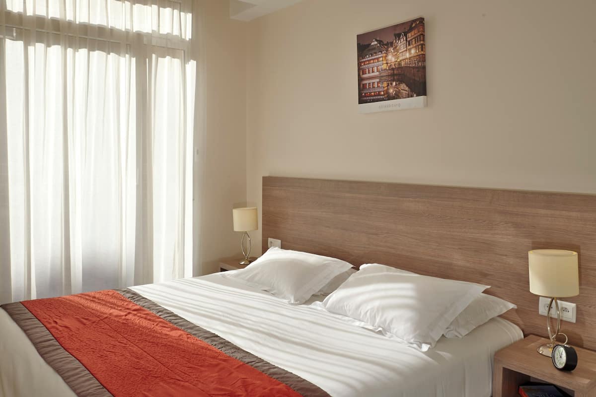 A hotel room with a large bed, two bedside tables with lamps, a nightstand with a book, and a cityscape painting on the wall.