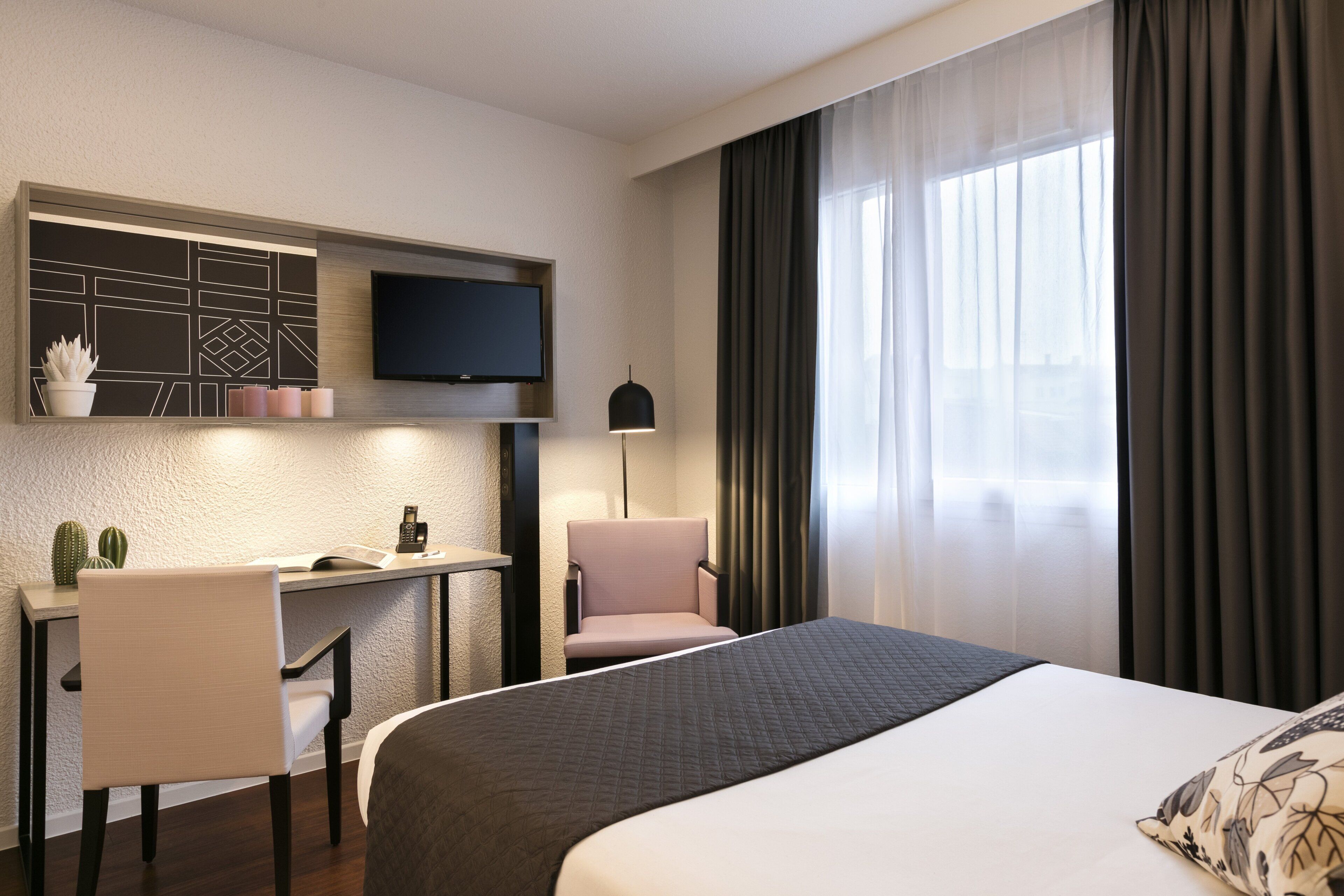Et hotelværelse med en seng, et skrivebord med stol, et fjernsyn og et vindue med gardiner.