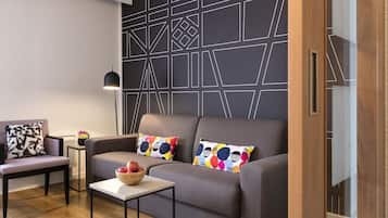 A modern living room with a grey sofa, colorful pillows, a small table with a bowl of fruit, and a geometric-patterned wall.