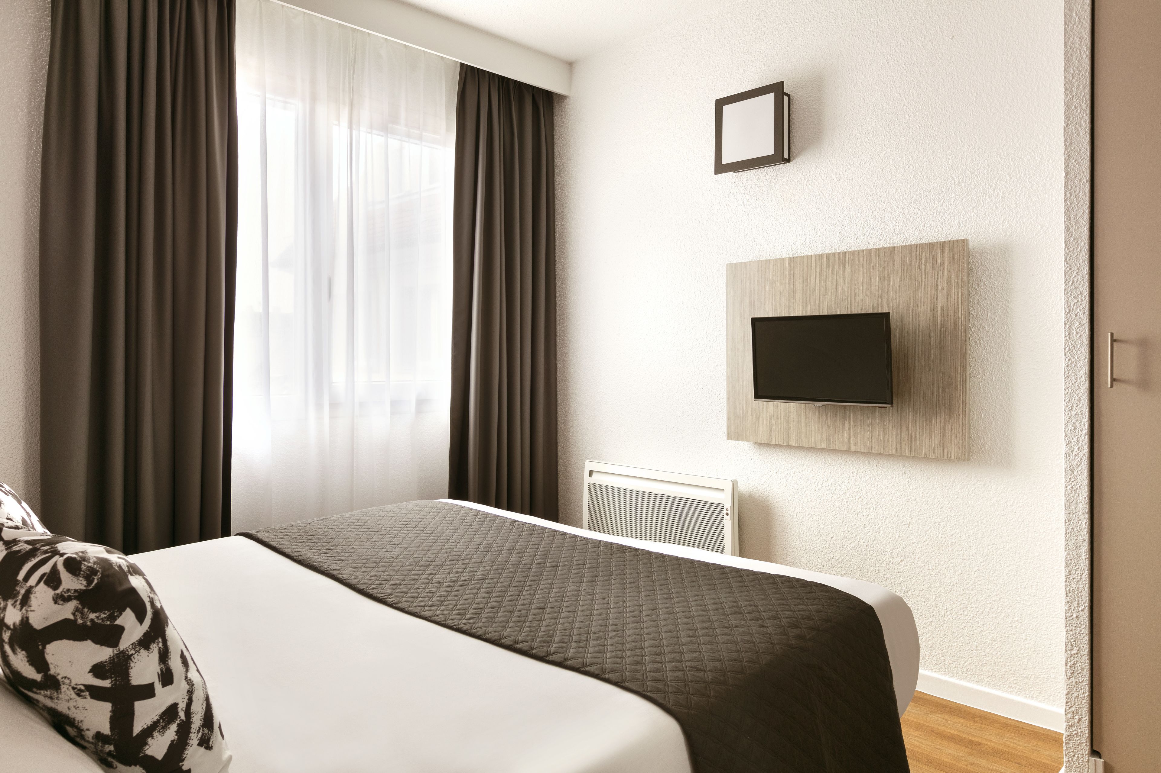 Une chambre d’hôtel avec un lit, un téléviseur fixé au mur et une fenêtre avec des rideaux.