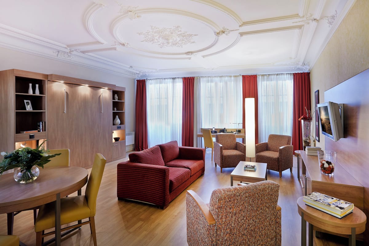 A modern hotel room with a sofa, armchairs, a dining table, and a TV.