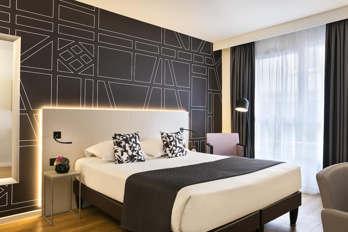Habitación de hotel moderna con una cama grande, una mesita de noche con lámpara y vistas a la ciudad a través de cortinas translúcidas.
