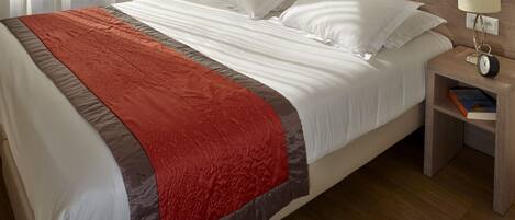 Una cama bien tendida con una colcha roja y gris, un cabecero de madera y una mesita de noche con una lámpara y un libro.