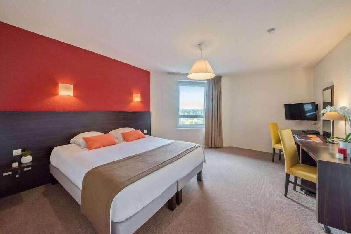 Ein Hotelzimmer mit einem großen Bett, einem Schreibtisch mit Stuhl, einem Fernseher und einem Fenster mit Vorhängen.
