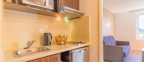 Cocina compacta con microondas, lavavajillas y pileta. Hay una cesta de frutas en la mesada y un sofá en el área de estar contigua.