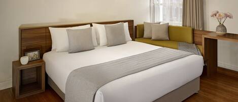 A hotel room with a large bed, bedside tables, a sofa, a nightstand, a vase with flowers, and a window with curtains.