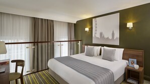 Habitació d'hotel amb un llit gran, una taula de treball amb una cadira, una tauleta de nit i una làmpada penjada a la paret.