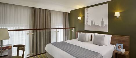 A hotel room with a large bed, a desk with a chair, a nightstand, and a wall-mounted lamp.